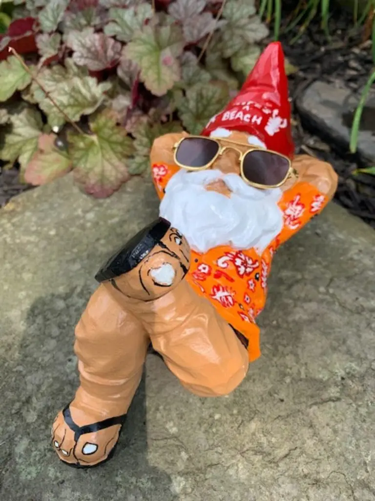 Garden Gnome with Life's A Beach written on red hat in white paint wearing sunglasses, flip flops, hands behind head, crossed leg, laying on back, relaxing, left