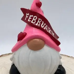 Birthday Gnome for February wearing pink hat with heart and lip shaped gems, and a dark red banner across that reads February in White, Front.