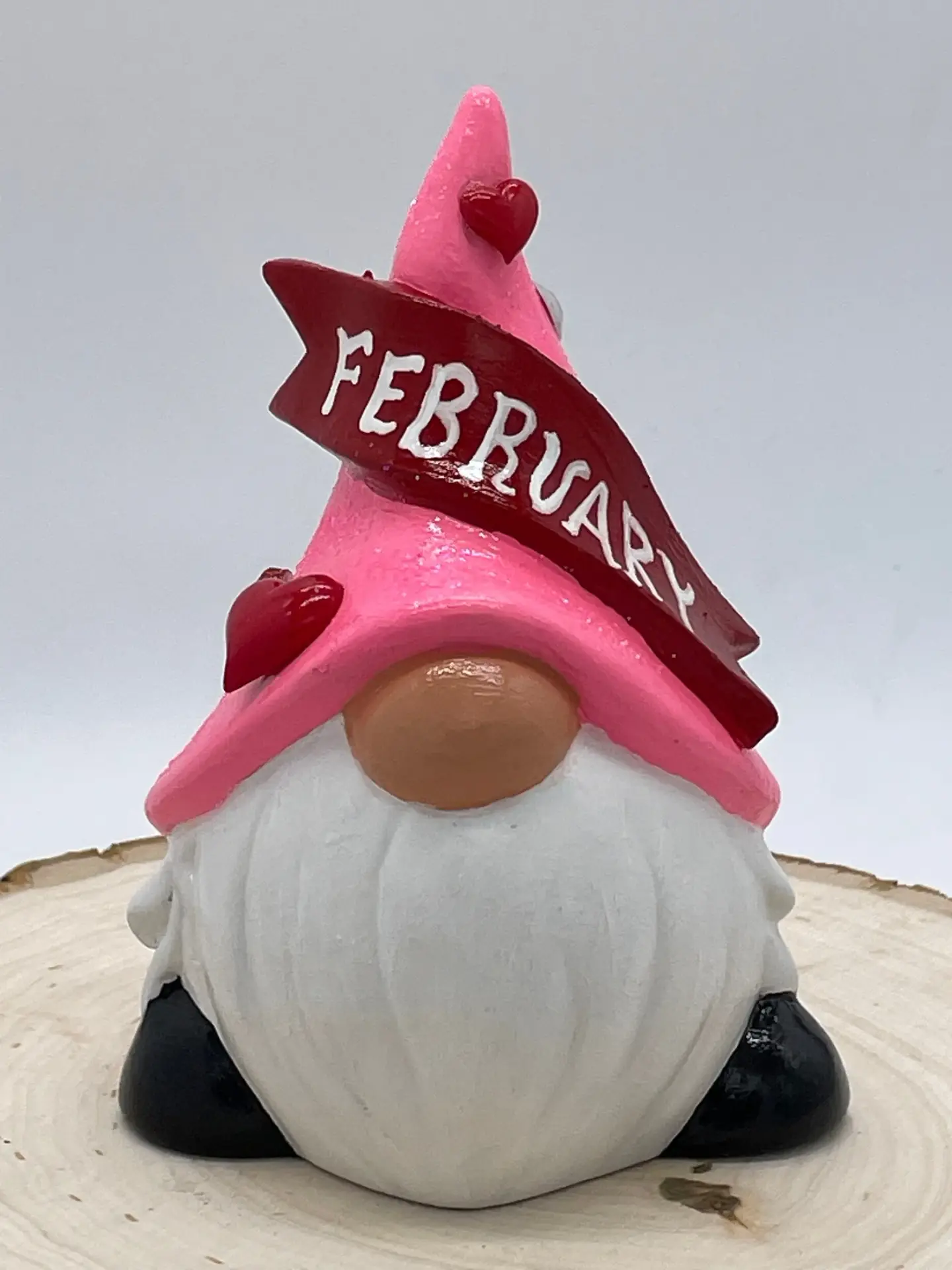 Birthday Gnome for February wearing pink hat with heart and lip shaped gems, and a dark red banner across that reads February in White, Front.