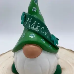 Birthday Gnome for March wearing green hat with white clovers, and a darker green banner across that reads March in White, Front