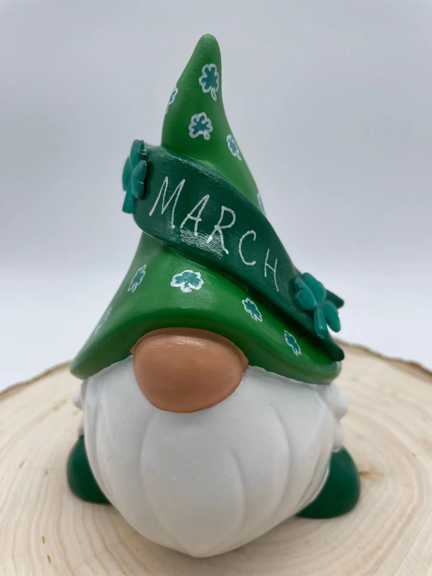 Birthday Gnome for March wearing green hat with white clovers, and a darker green banner across that reads March in White, Front